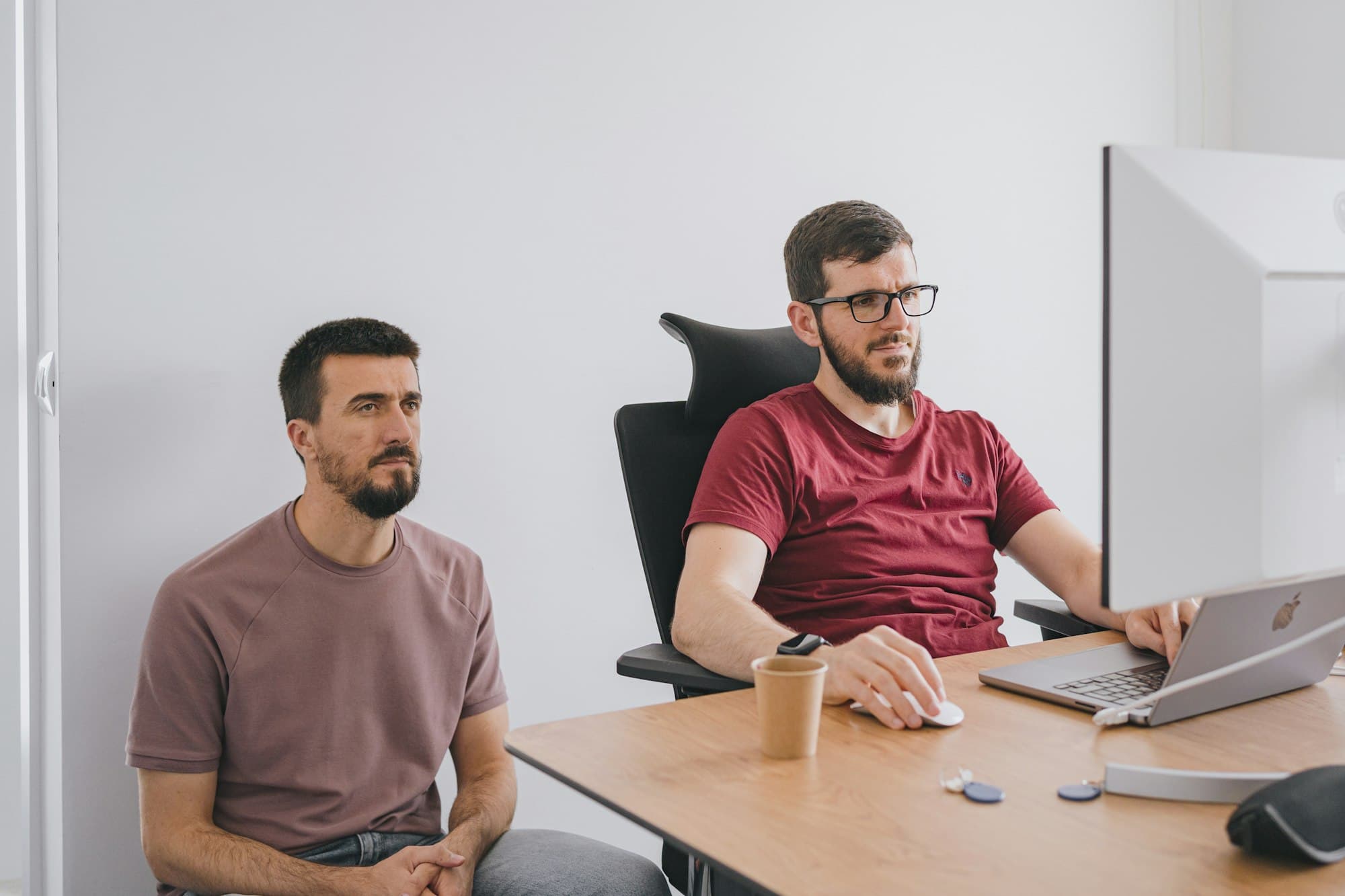 Two coworkers in a small office, one with arms crossed, both looking serious